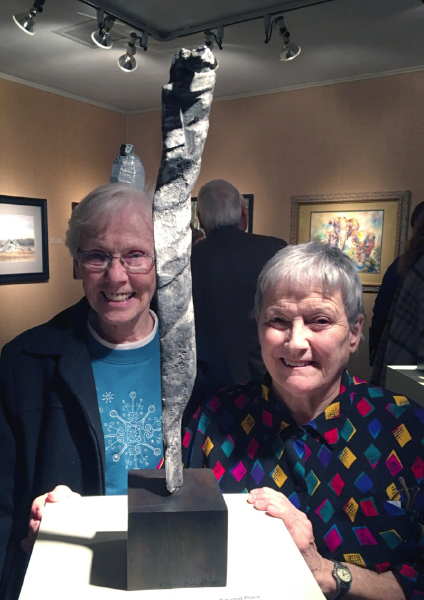 Two women smiling with a small silver sculpture at a gallery event inside.