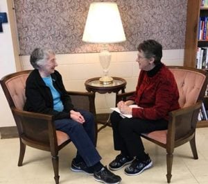Two short-haired women sitting together and conversing in a sitting nook next to a book shelf inside