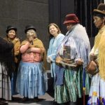 Six diverse actresses dressed in large costume dresses and hats, conversing.