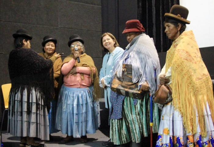 Six diverse actresses dressed in large costume dresses and hats, conversing.