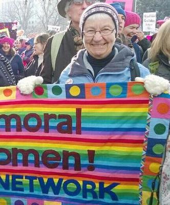 Woman in glasses, hat, and gloves smiling holding up a colorful sign at the womens march