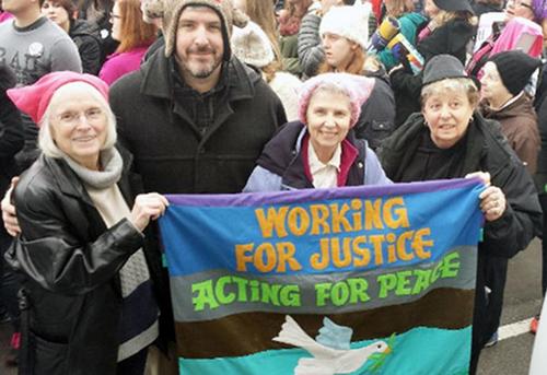 Four individuals smiling for a picture displaying a banner that says: Working for Justice Acting for Piece in a crowded street at the womens march.