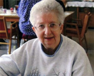 A short-haired with glasses woman smiling in a grey sweater at a table indoors.
