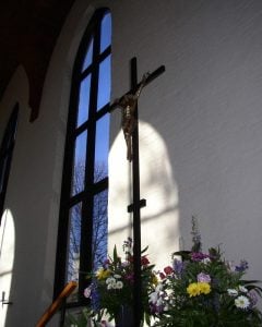 Light from an arched window illuminates a tall crucifix in a church. Spring flower arrangements surround the base of the cross.