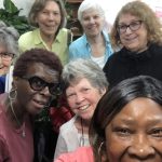 Seven diverse women smile together for a group selfie inside.