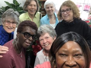 Seven diverse women smile together for a group selfie inside.