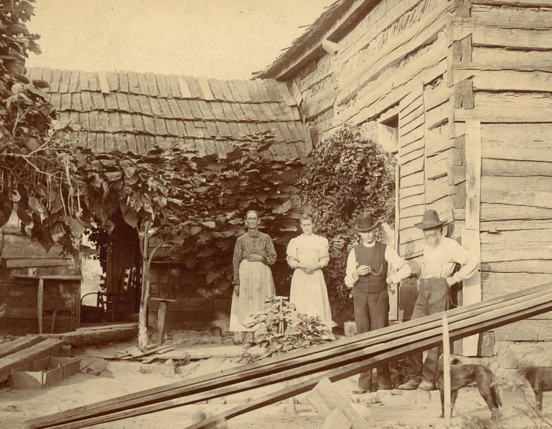 Archival photo of two women in dresses and aprons and two men in hats standing in front of a wooden house.