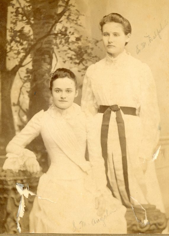 Archival sepia photo of two young women posing for a photo - one sitting, one standing.