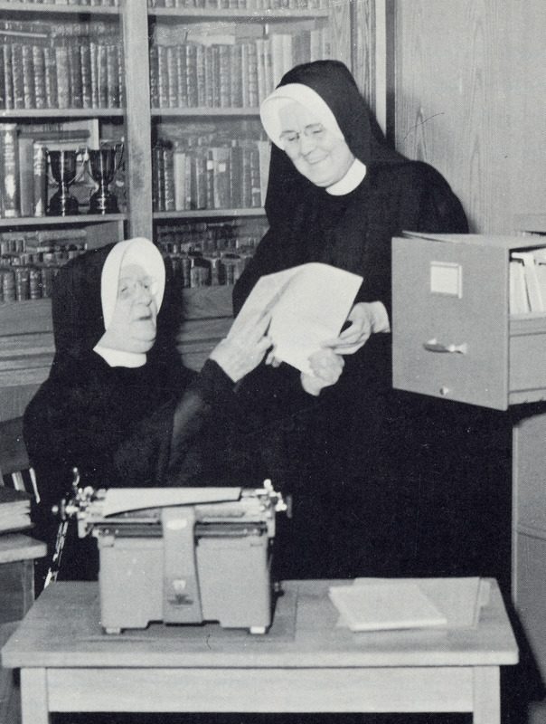 Archival photo of two nuns in habits looking over a sheet of paper they've pulled from a typewriter. A file cabinet drawer stands open.