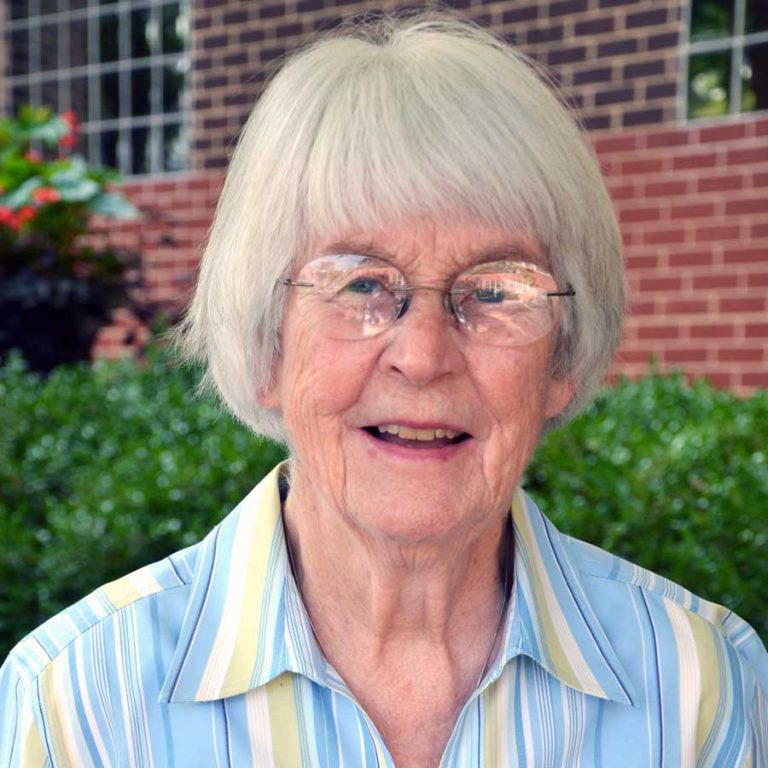 A woman with short grey hair with bangs and round glasses wearing a yellow and blue striped collared button-up shirt smiling for a headshot picture outdoors in front of a red brick building and green bush.