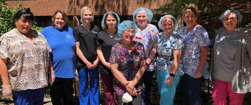 Nine women in scrubs, some wearing hairnets, pose for a photo in the courtyard.