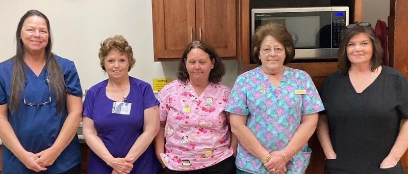 Five women in colorful scrubs line up for a photo in a staff kitchen.