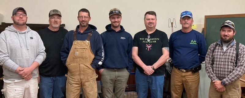 Seven men in casual work clothes (ball caps, overalls, sweatshirts and canvas pants) line up for a group photo.