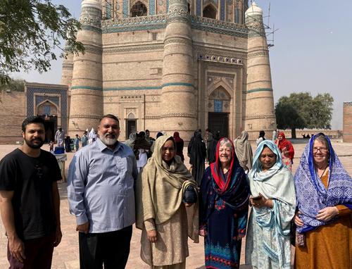 Six individuals smile in front of a beige and blue monument. It is sunny outside, and the four women are wearing head scarves.