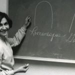 A woman turns from the chalkboard where she has drawn several figures to smile for the camera.