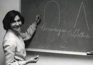 A woman turns from the chalkboard where she has drawn several figures to smile for the camera.