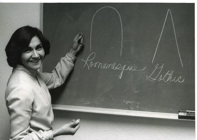 A woman turns from the chalkboard where she has drawn several figures to smile for the camera.