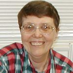A woman with short brown hair and oval-shaped glasses wearing a red and blue plaid collared shirt smiling brightly for a headshot picture indoors in front of a window closed with white blinds.