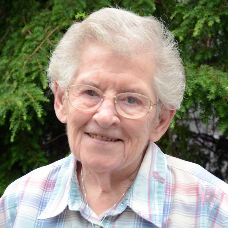 A woman with short white hair and round glasses wearing a light-colored plaid collared button-up shirt smiling for a headshot picture outdoors in front of pine tree greenery.