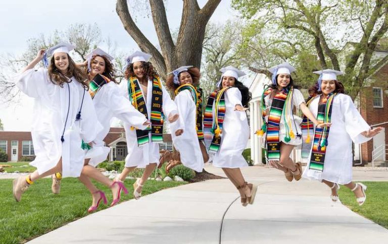 Students in white graduation caps and gowns, and colorful stoles and tassels leap together for joy!