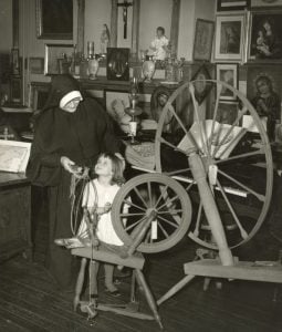 Black and white photo of a nun in habit looking down at a young girl. They stand next to a spinning wheel in a room surrounded by Christian art and artifacts.