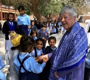 A short-haired woman in a blue shall stands holding hands with young school children outside on a sunny day.