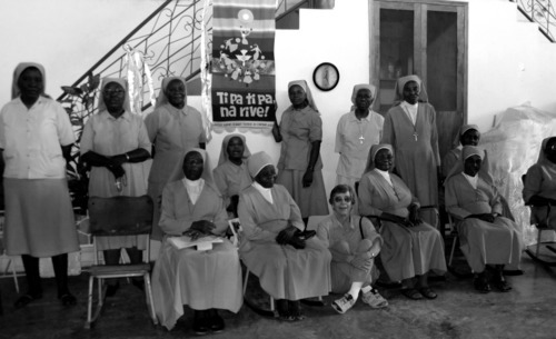 A black and white image of a large group of people in nuns' habits smiling around a banner in a house that reads, "Ti pa, ti pa na rive!" (Little by little, we will continue.)