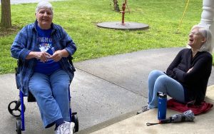 Two women sitting down laughing and conversing on a sidewalk next to green grass on a sunny day.