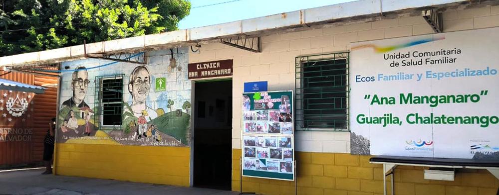Outside of a building with a mural of Oscar Romero and Ann Manganaro on the left and the name "'Ann Manganaro' Guarjila, Chaletenango" on the right