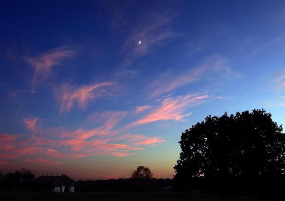 Clouds glow pink and orange in a navy sky, as a house and trees are silhouetted against the fading glow of dusk.