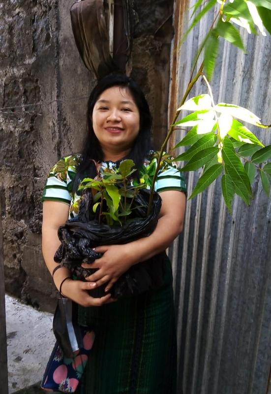A woman stands outside next to a wall holding a pot with a young tree in it.