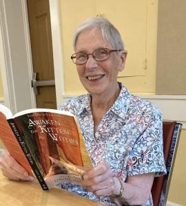 A woman with short hair and glasses sits at a table with an open book in her hands.