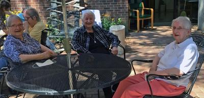 Three short-haired women mingle at an outdoor table under an umbrella on a sunny day.