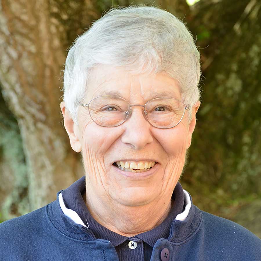 A woman with short white hair and round wire-framed glasses wearing a navy blue collared shirt and a navy blue jacket with a white collar trim smiling brightly for a headshot picture outdoors in front of a large treetrunk.