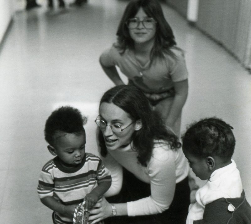 Archival photo of a woman kneeling down in a hallway with three small children around her. 