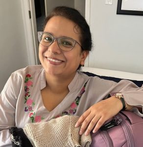 Woman in an embroidered blouse and glasses smiles brightly while leaning on a suitcase inside.
