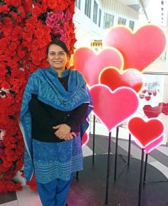 A woman wearing a blue shawl and skirt smiling in front of some pink and red heart posters and a red rose wall.