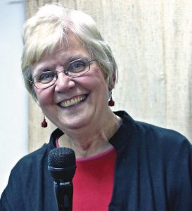 Short haired woman wearing glasses, a red shirt and navy blue jacket smiles for a picture while using a microphone.