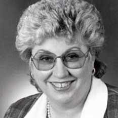 A black and white headshot picture of a woman smiling with short hair and large round glasses wearing a plaid blazer with a white collar, hoop earrings and a pearl necklace.
