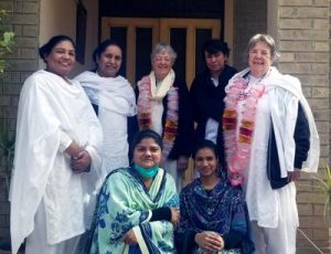 Seven diverse women in shawls and scarves smile together for a group photo in front of a building’s front door.