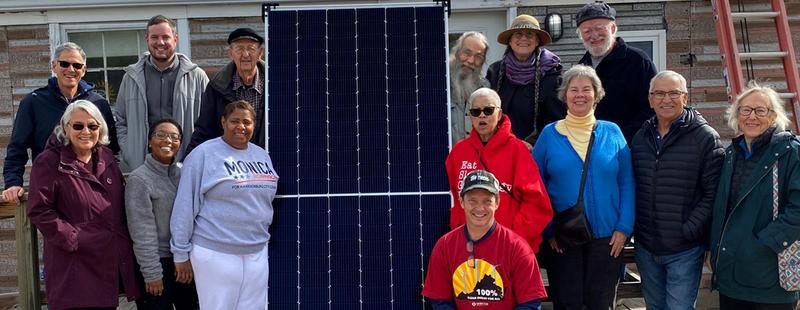 A diverse group of people gather around a solar panel leaning against a building on a chilly morning.