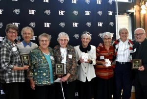 Eight diverse, well dressed individuals smile together in front of a backdrop, while holding three plaques and two small white statues.