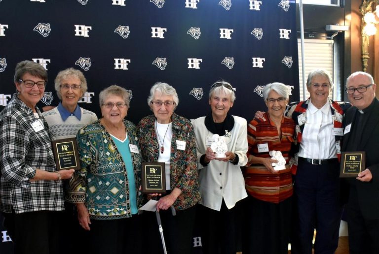 Eight diverse, well dressed individuals smile together in front of a backdrop, while holding three plaques and two small white statues.