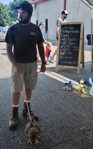 A man wearing a baseball cap, t-shirt, and khaki shorts walking a black and white speckled chicken on a leash.