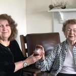 Two women smile together for a picture while holding a jar of jam together.
