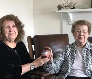 Two women smile together for a picture while holding a jar of jam together.