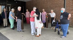 Long black haired woman wearing a backpack is circled and hugged by friends saying goodbye outside of a brick building.