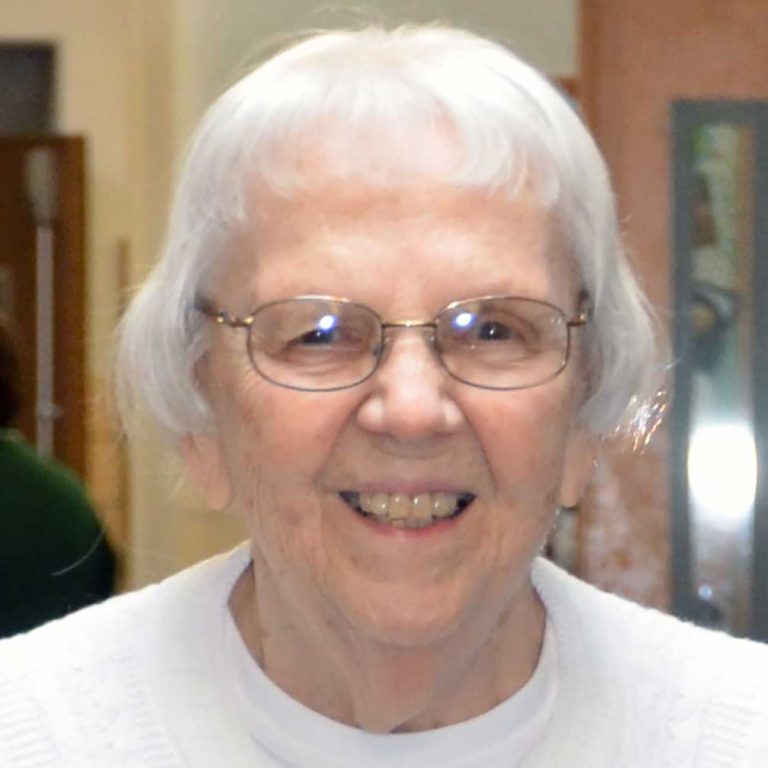 A woman with short white hair and wire-framed oval-shaped glasses wearing a plain white shirt smiling brightly for a headshot picture indoors.