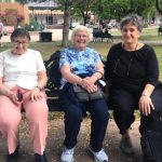 Three women sit together on a bench in a park smiling together for a picture.