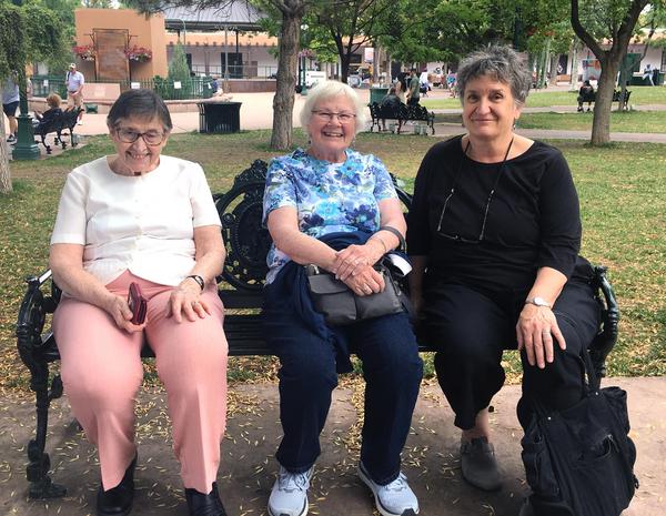 Three women sit together on a bench in a park smiling together for a picture.